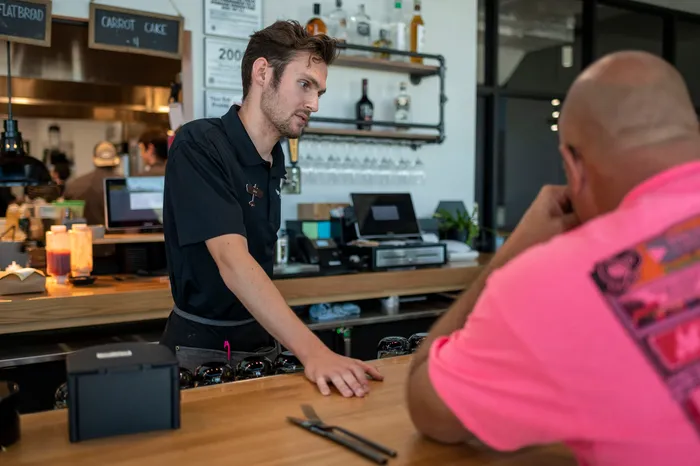 A bartender in a black polo talking with a customer in a pink shirt at a wooden bar
