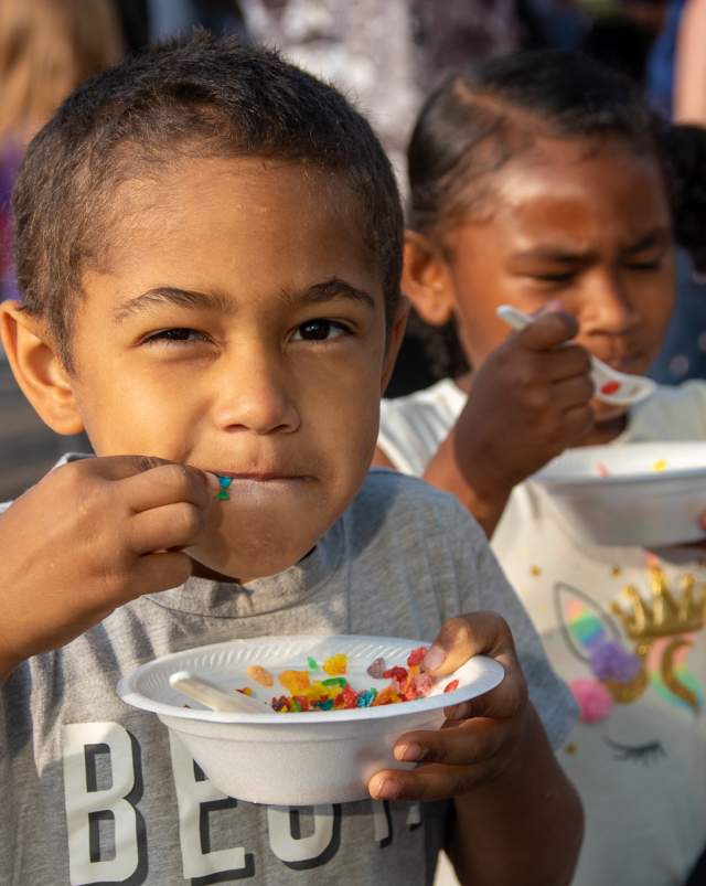 A young boy eating colorful cereal from a paper bowl at the National Cereal Festival, with another child behind him