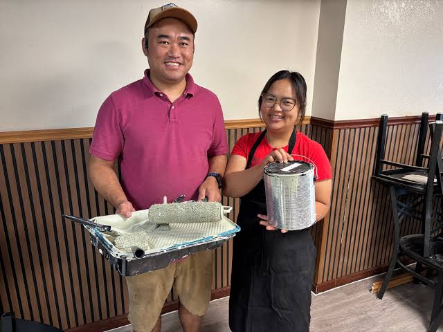 Two smiling business owners holding a paint roller tray and a can of paint inside their restaurant