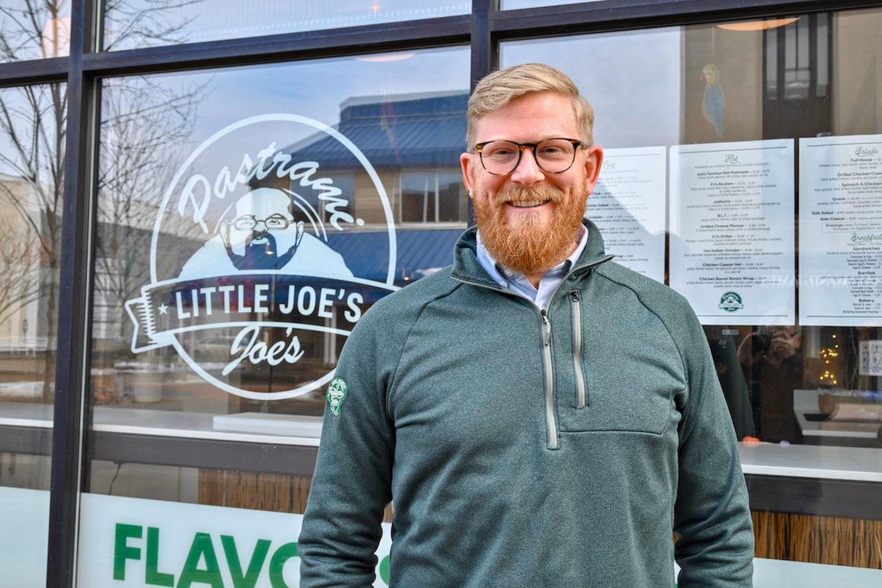 A smiling business owner in a green quarter-zip standing outside the Pastrami Little Joe’s storefront