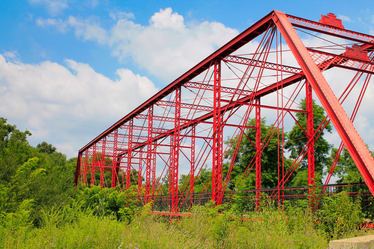 A red iron truss bridge surrounded by greenery under a blue sky