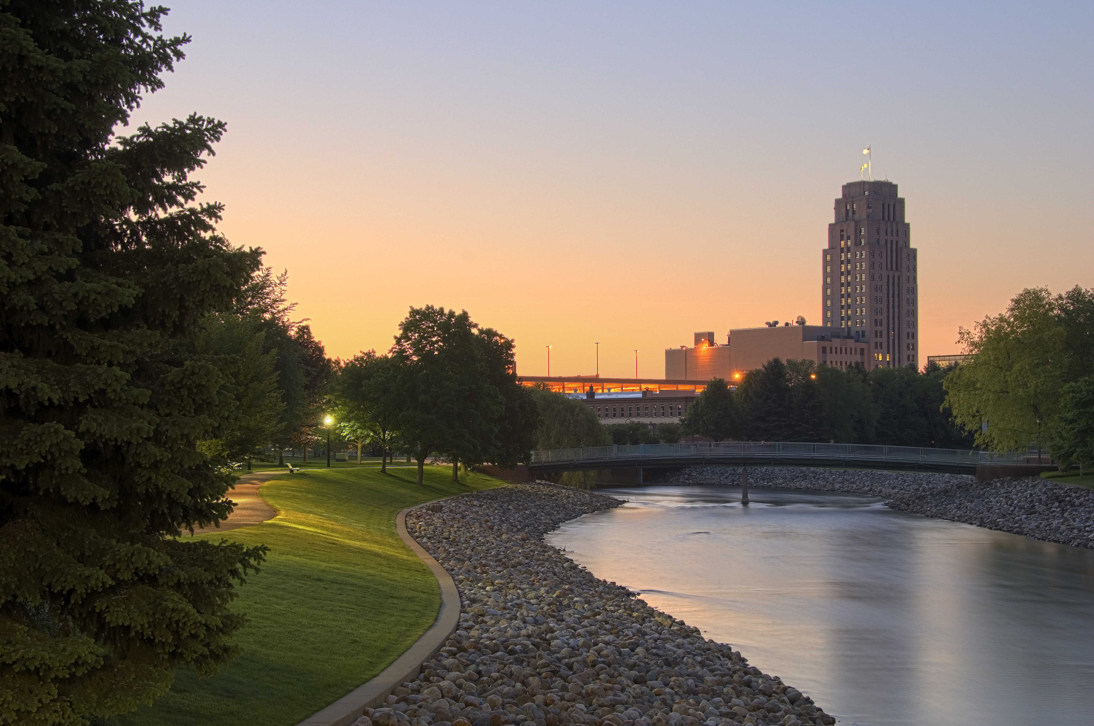 A riverside walking path and footbridge in Battle Creek at dusk, with the tower glowing in the distance