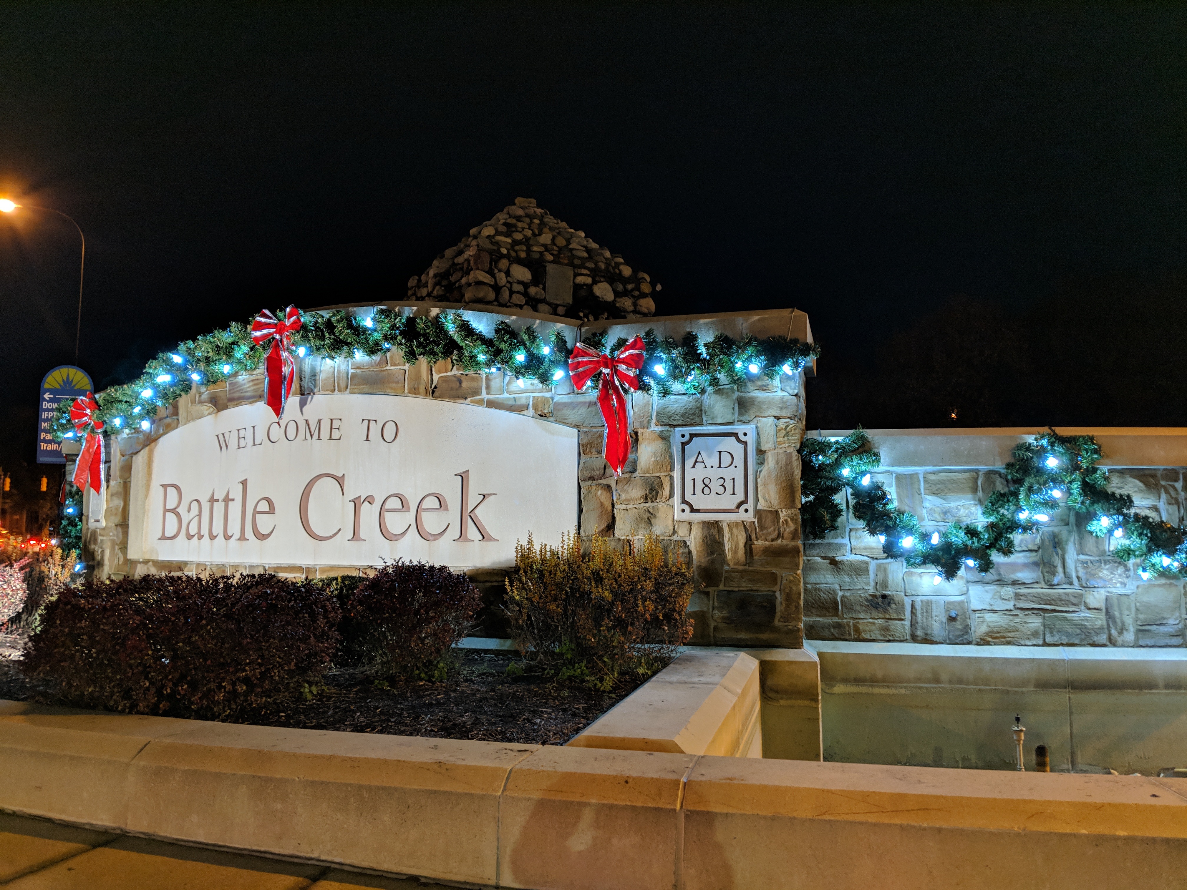 The Battle Creek welcome sign at night, decorated with greenery, lights, and red bows